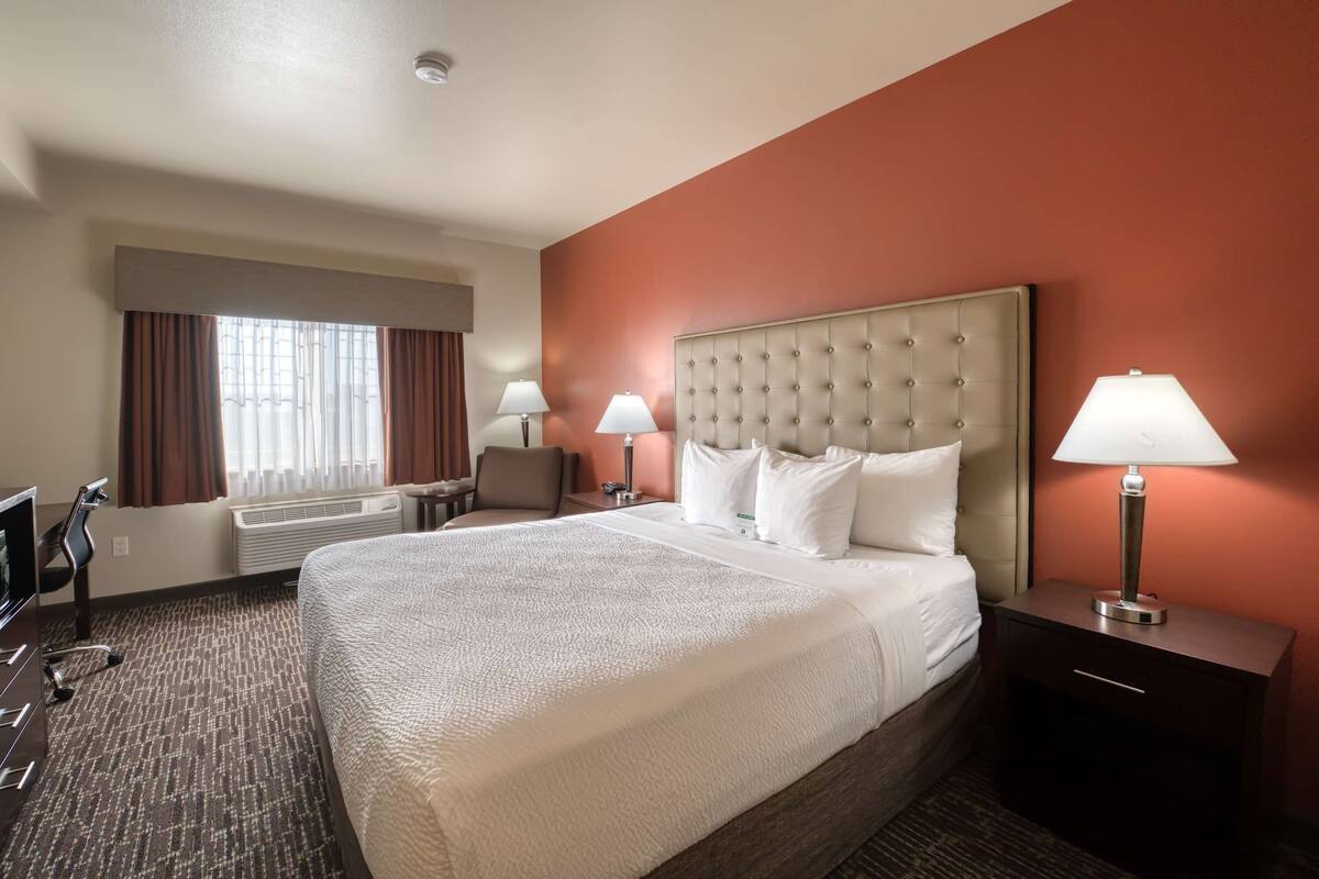 A hotel room with a large bed, two bedside lamps, a desk with a chair, and a window with curtains.