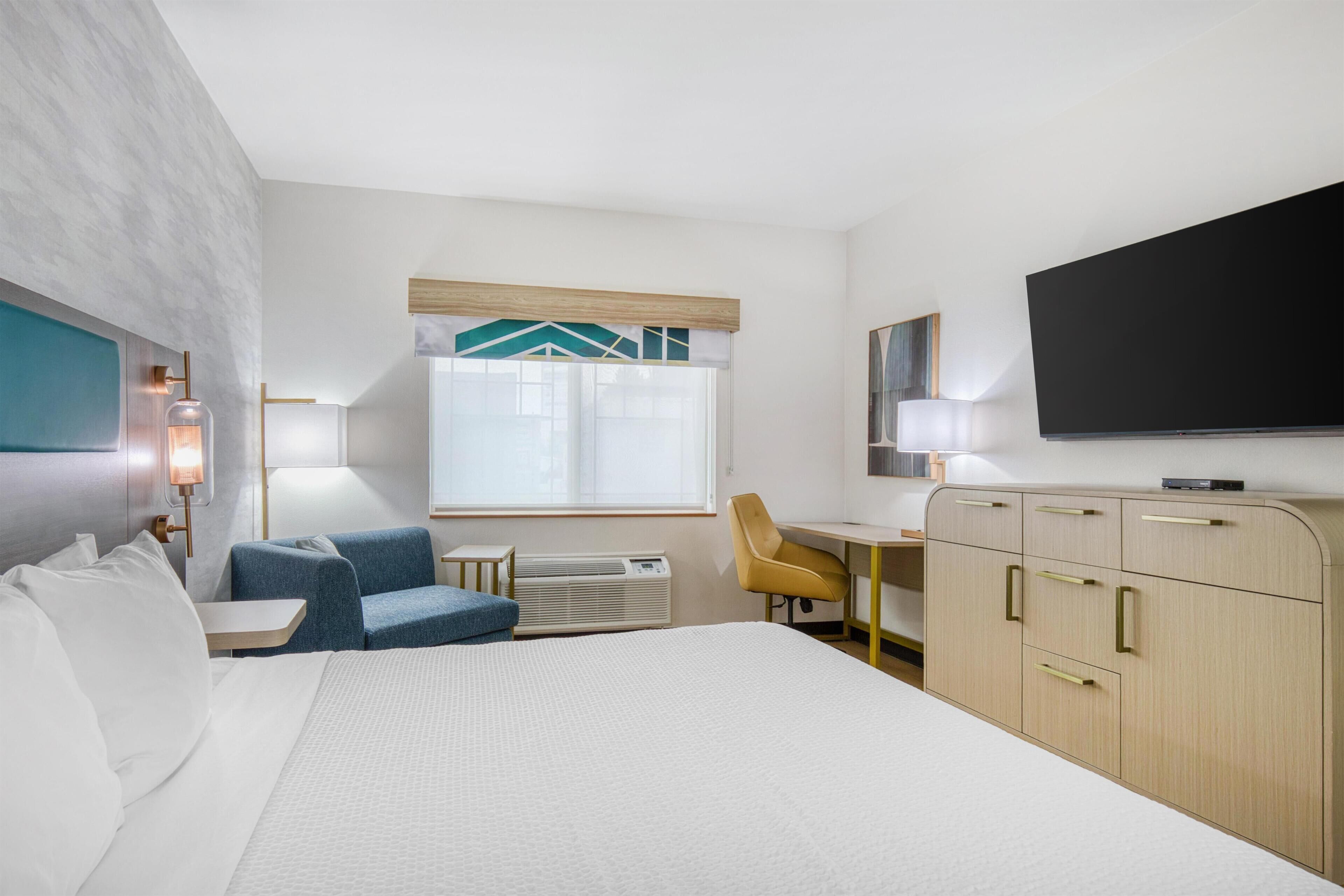 Une chambre d’hôtel avec un lit, un bureau, une chaise, une télévision et une fenêtre avec des rideaux.
