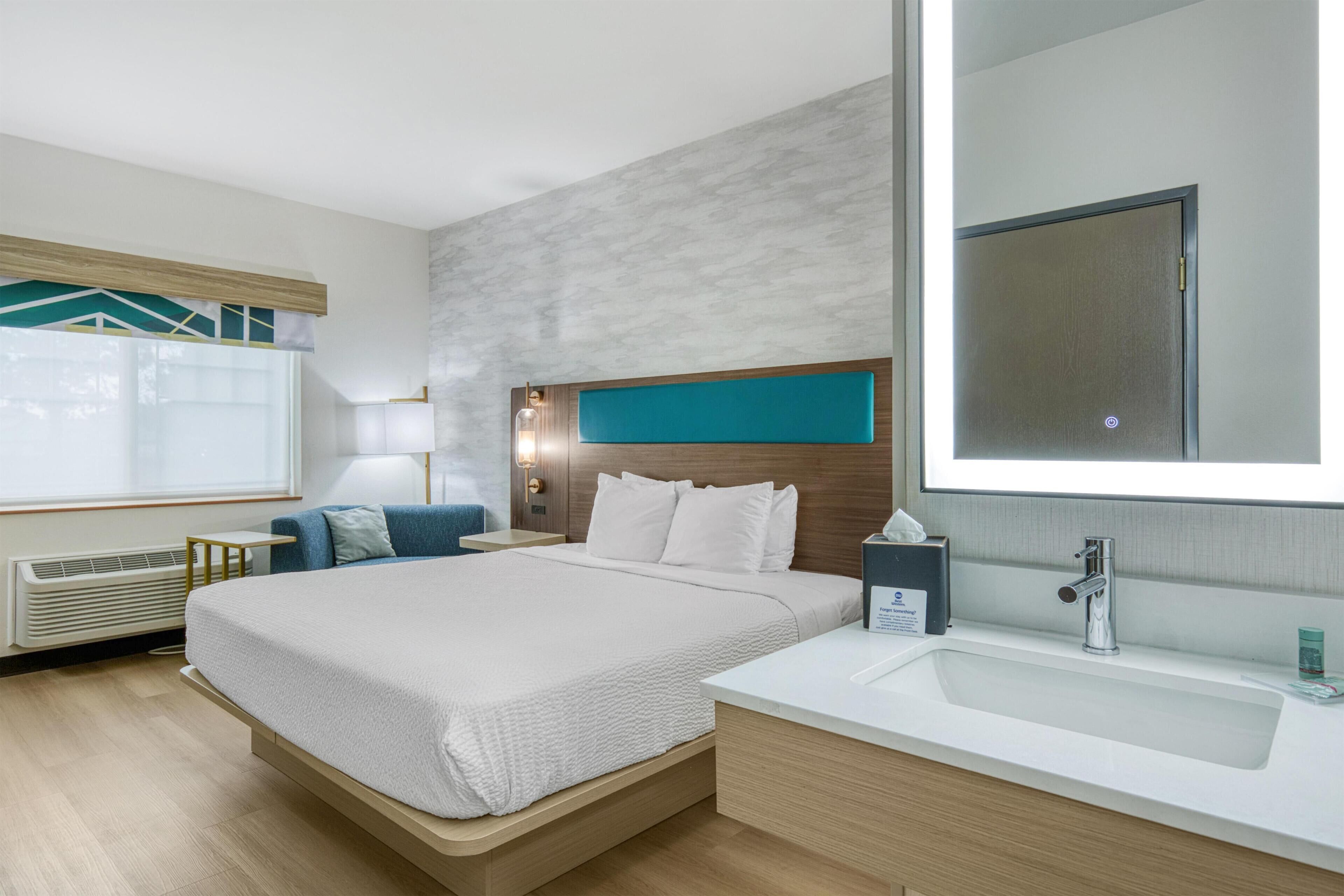 A hotel room with a large bed, a bedside table, a nightstand, a mirror, a window with blinds, and a bathroom with a sink and mirror.