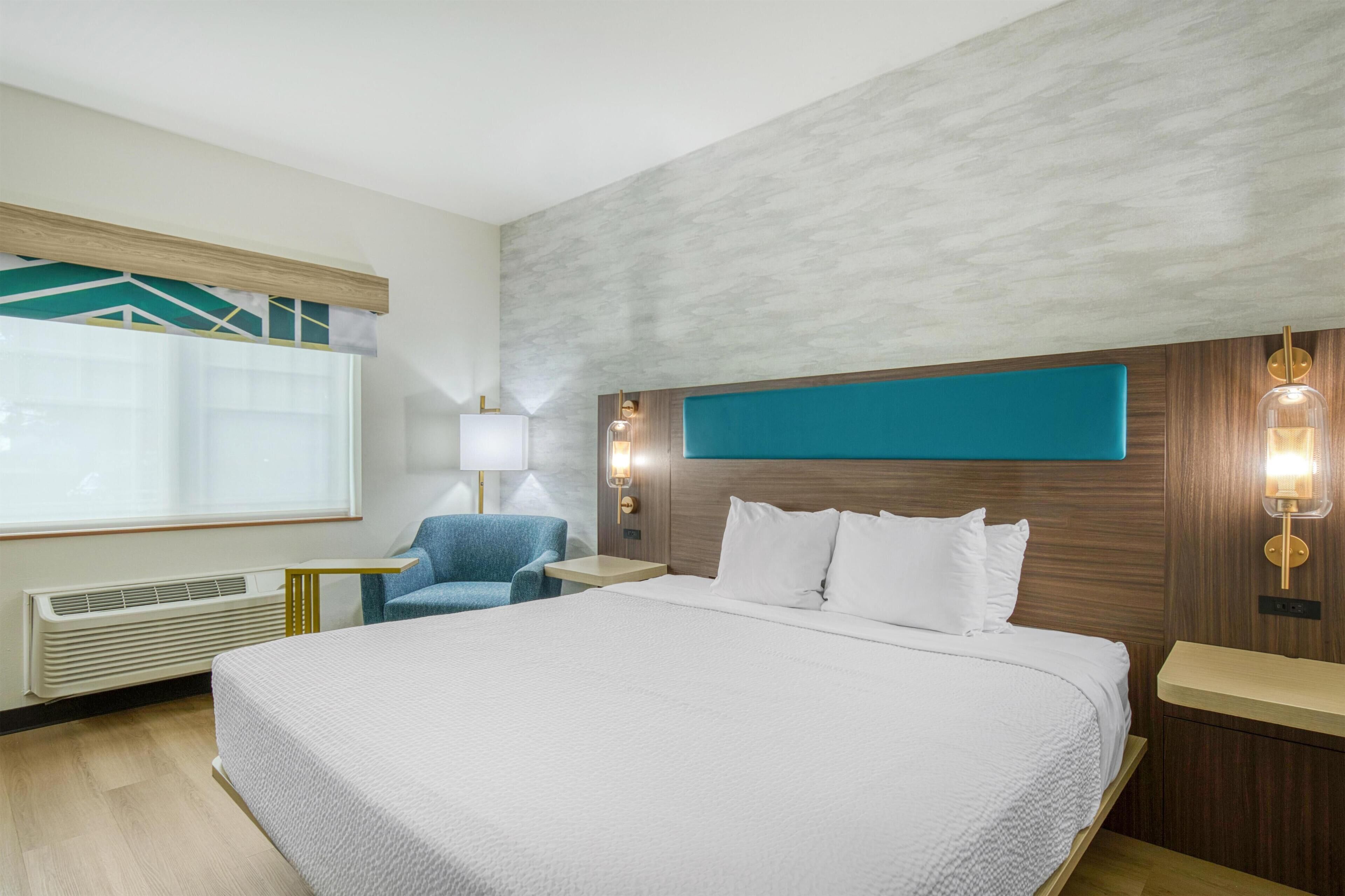A hotel room with a large bed, a side table, a chair, a lamp, and a view of the outdoors.