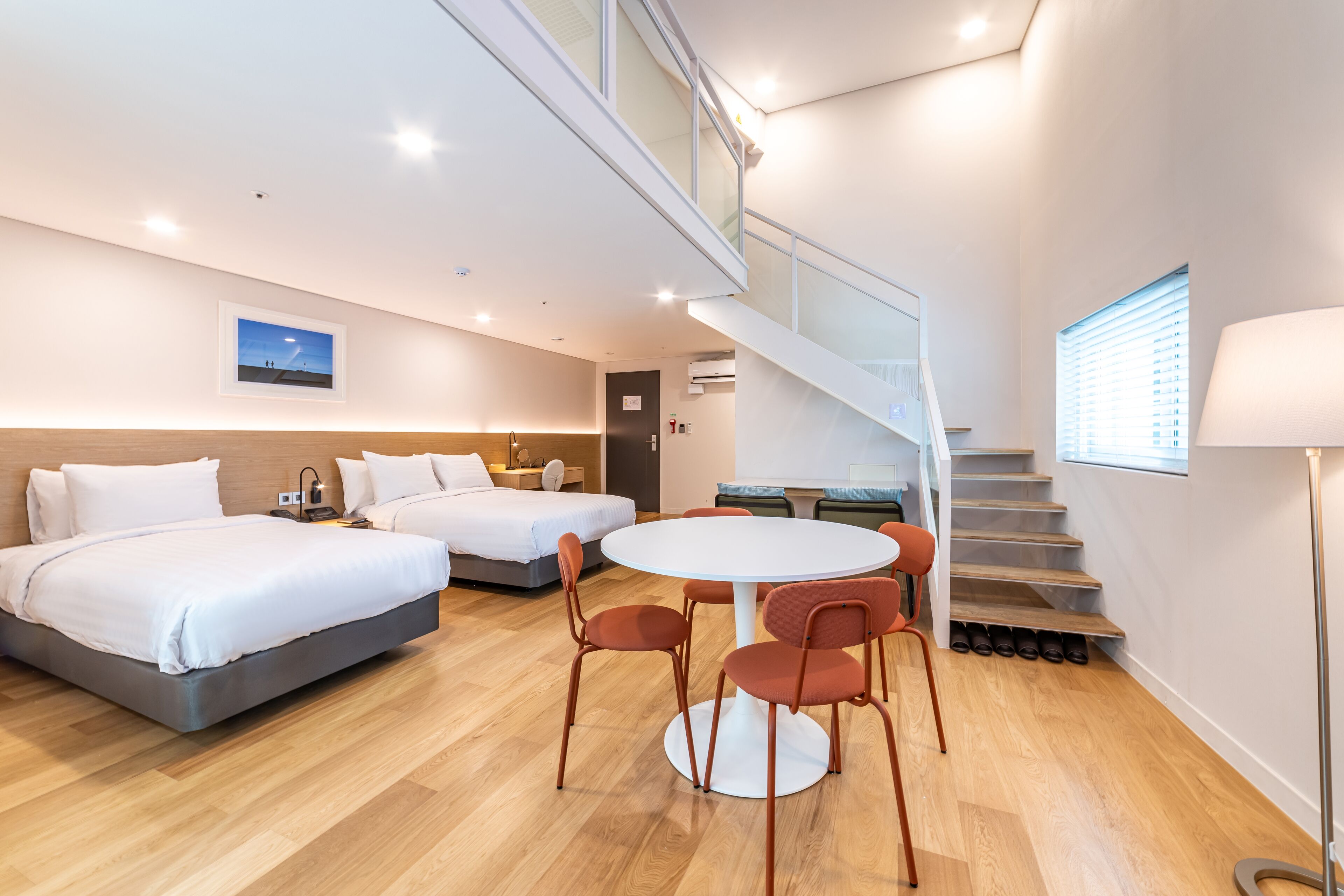 A modern hotel room with two beds, a round dining table with chairs, a staircase, and a view of the sky.