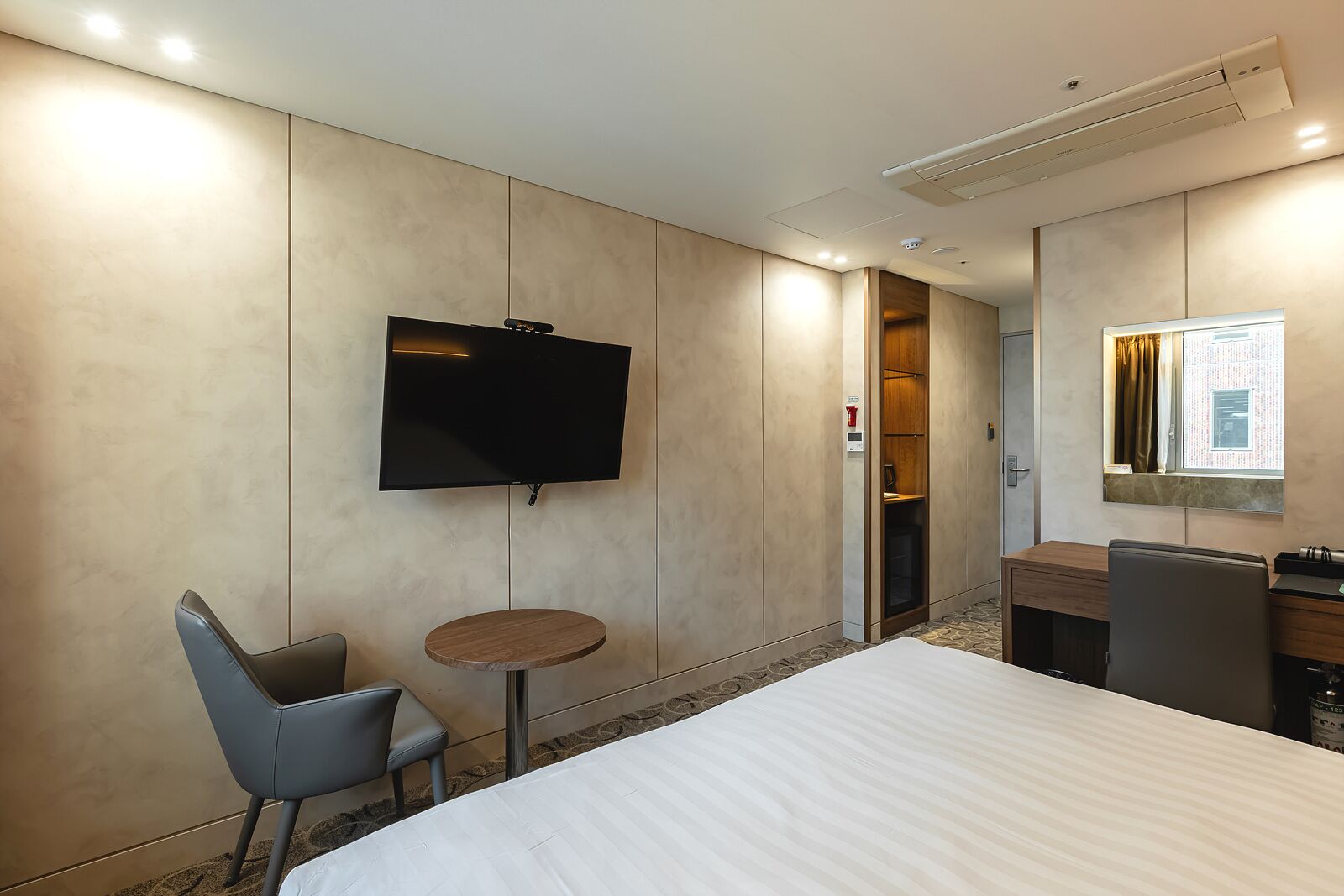 A hotel room with a bed, a TV mounted on the wall, a small round table, a chair, a desk with a chair, and a window with curtains.