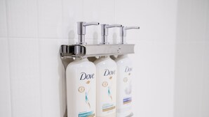 Tre Dove shampoo flasker på en hvid hylde foran en hvid flisevæg.