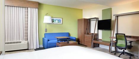 A hotel room with a bed, a desk, a chair, a TV, a sofa, and a window with curtains.