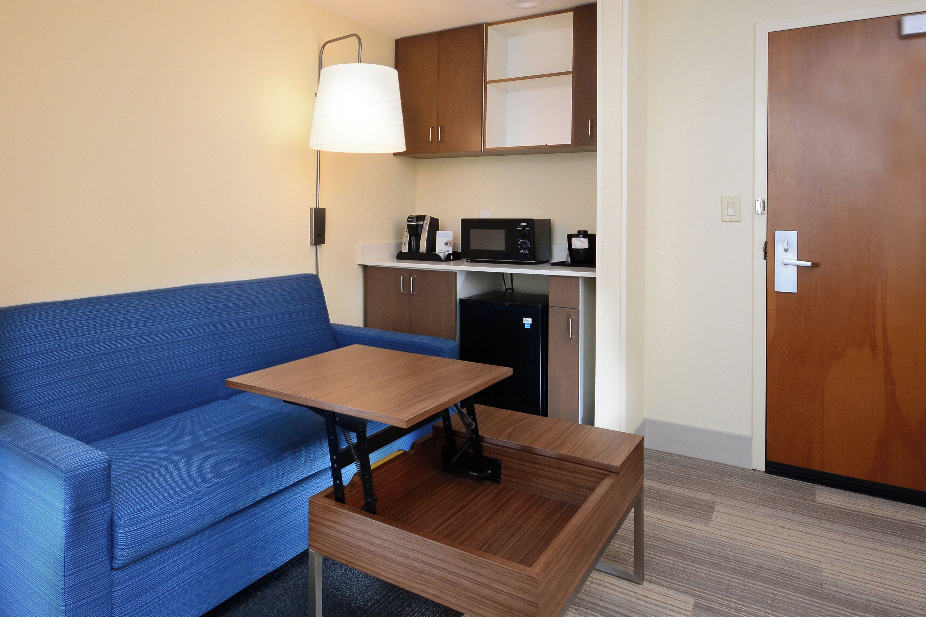 Habitació compacta amb un sofà blau, una taula de fusta petita, un microones, una cafetera i un armari.