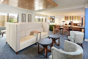 Un hall d’hôtel moderne avec un canapé d’angle, des tables rondes et un espace bar.