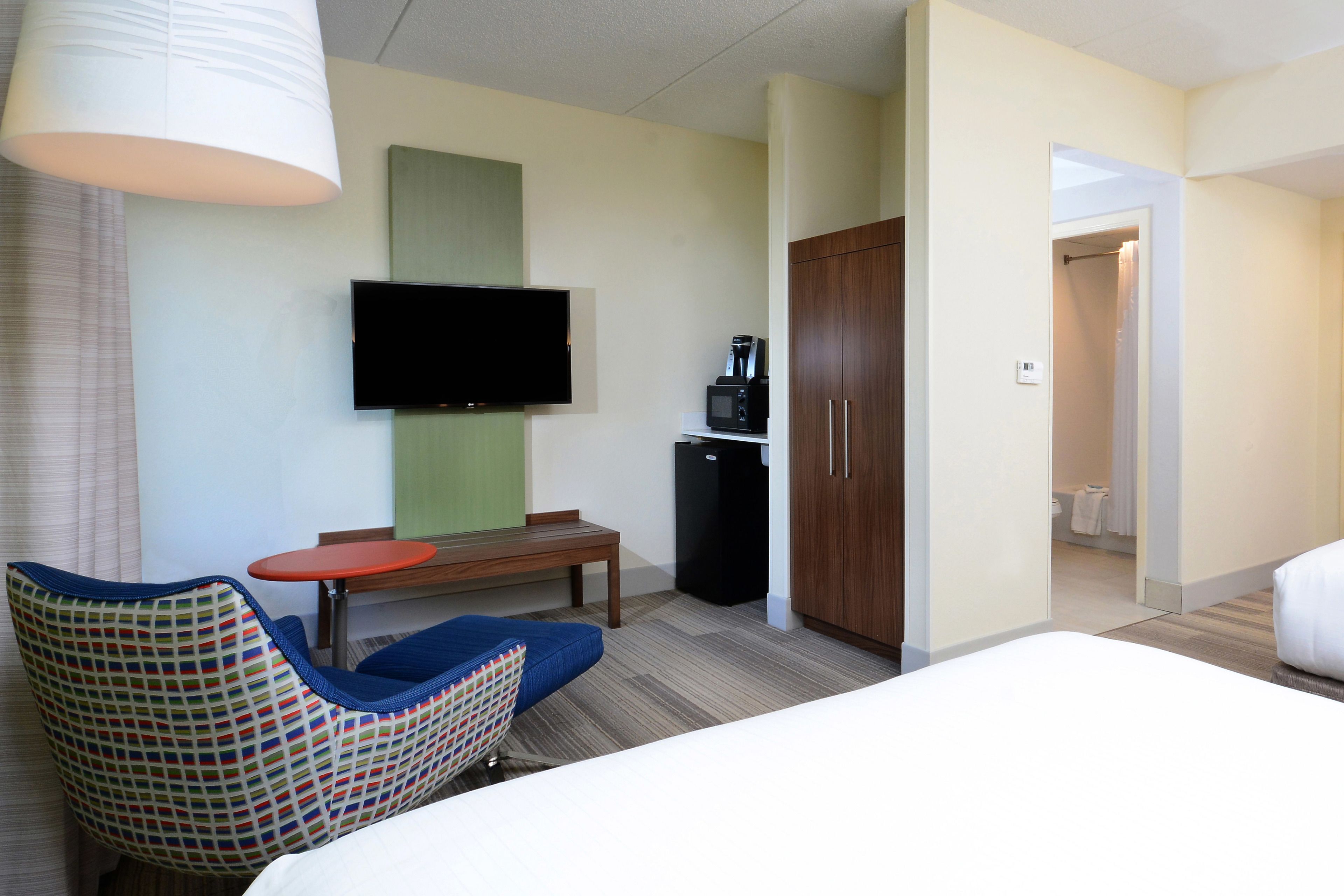 A hotel room with a bed, a TV mounted on the wall, a small table, a chair, a microwave, and a wardrobe.
