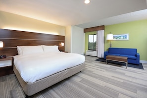 A hotel room with a large bed, a blue sofa, a small table, a nightstand, a lamp, and a window with curtains. - Holiday Inn Express & Suites Raleigh Durham Airport at RTP by IHG (Durham)