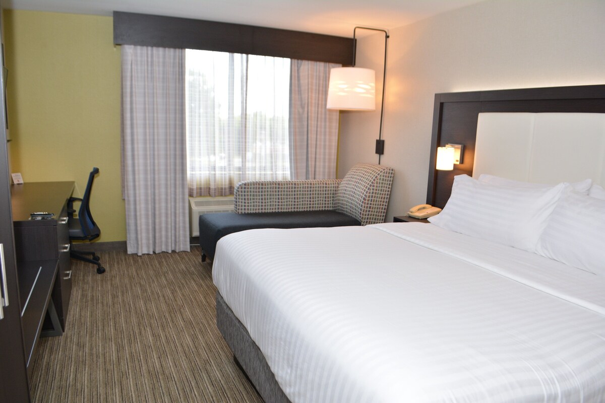 A hotel room with a large bed, a desk, a chair, a lamp, and a window with curtains.