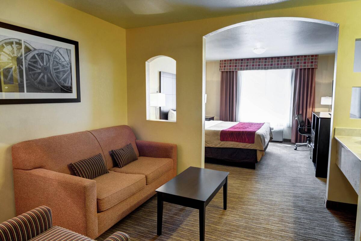 Una habitación de hotel con sofá, una mesita, una cama con una manta roja, un escritorio y un espejo.