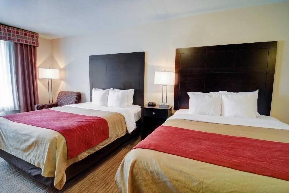Habitación de hotel con dos camas, cada una con cabecera, mesitas de noche, lámparas y una colcha roja.
