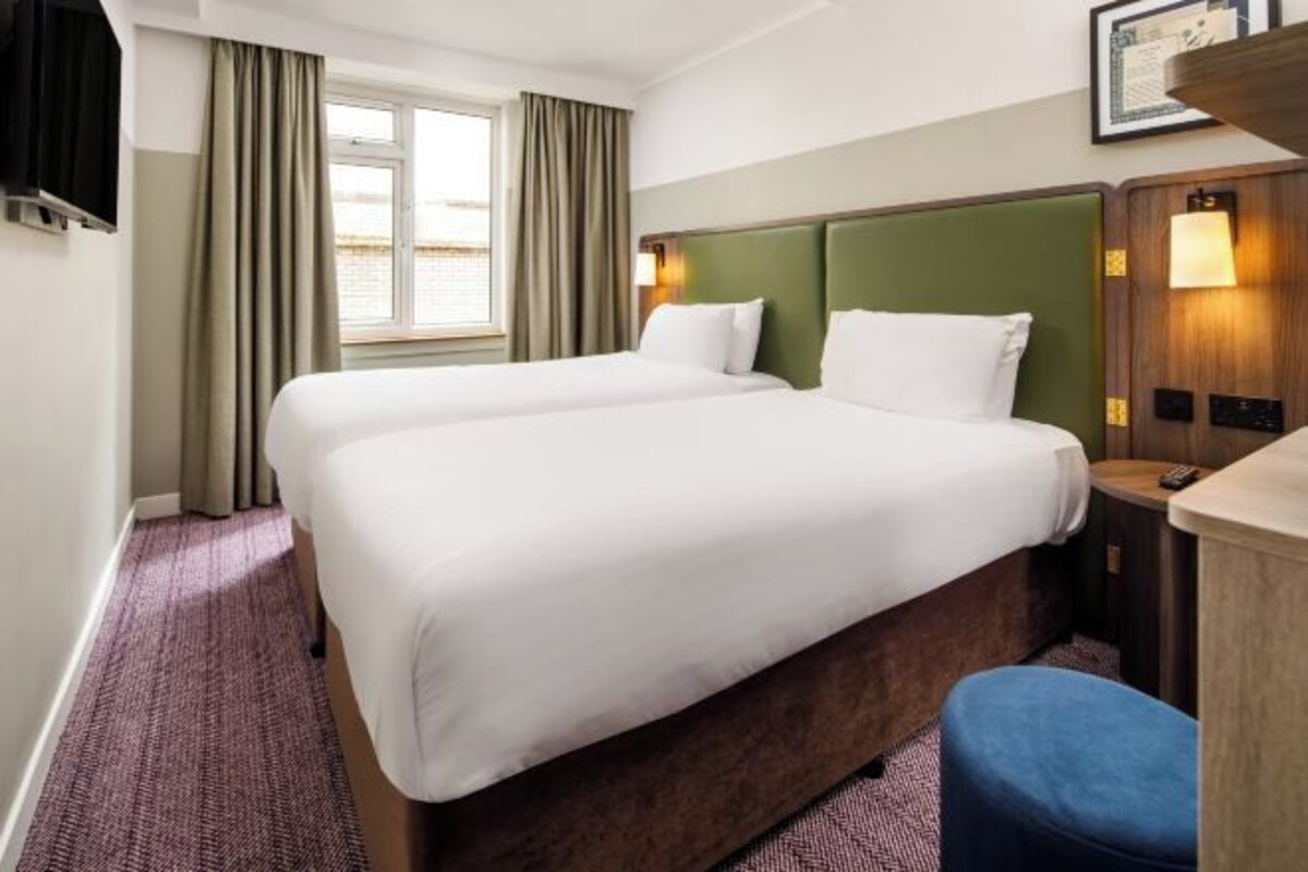 A hotel room with two beds, a window with curtains, a small table with a lamp, and a wall-mounted TV.