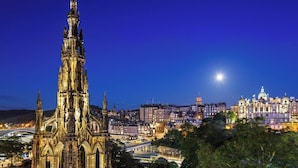 Una catedral de estilo gótico con intrincadas agujas y una imponente torre del reloj, rodeada por un paisaje urbano con edificios históricos y un puente.