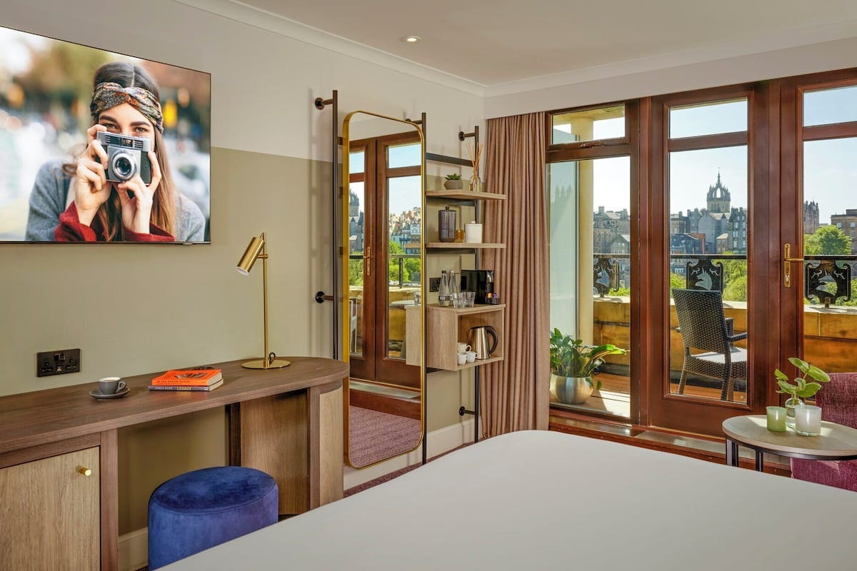 Habitación de hotel con cama, escritorio con lámpara, estantería, vista a la ciudad y televisor en la pared.
