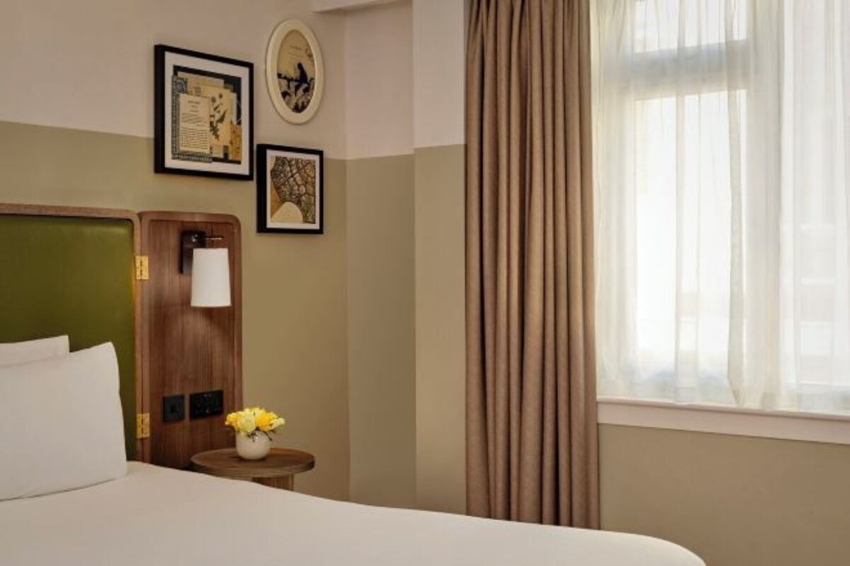 A hotel room with a bed, a nightstand with a lamp and flowers, a window with curtains, and framed pictures on the wall.