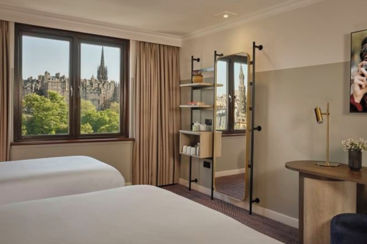A hotel room with two beds, a city view, a mirror, a shelf with toiletries, and a desk with a lamp.