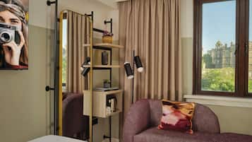 A hotel room with a sofa, a coffee table, a mirror, a shelf with decorative items, and a window with a view of a building.