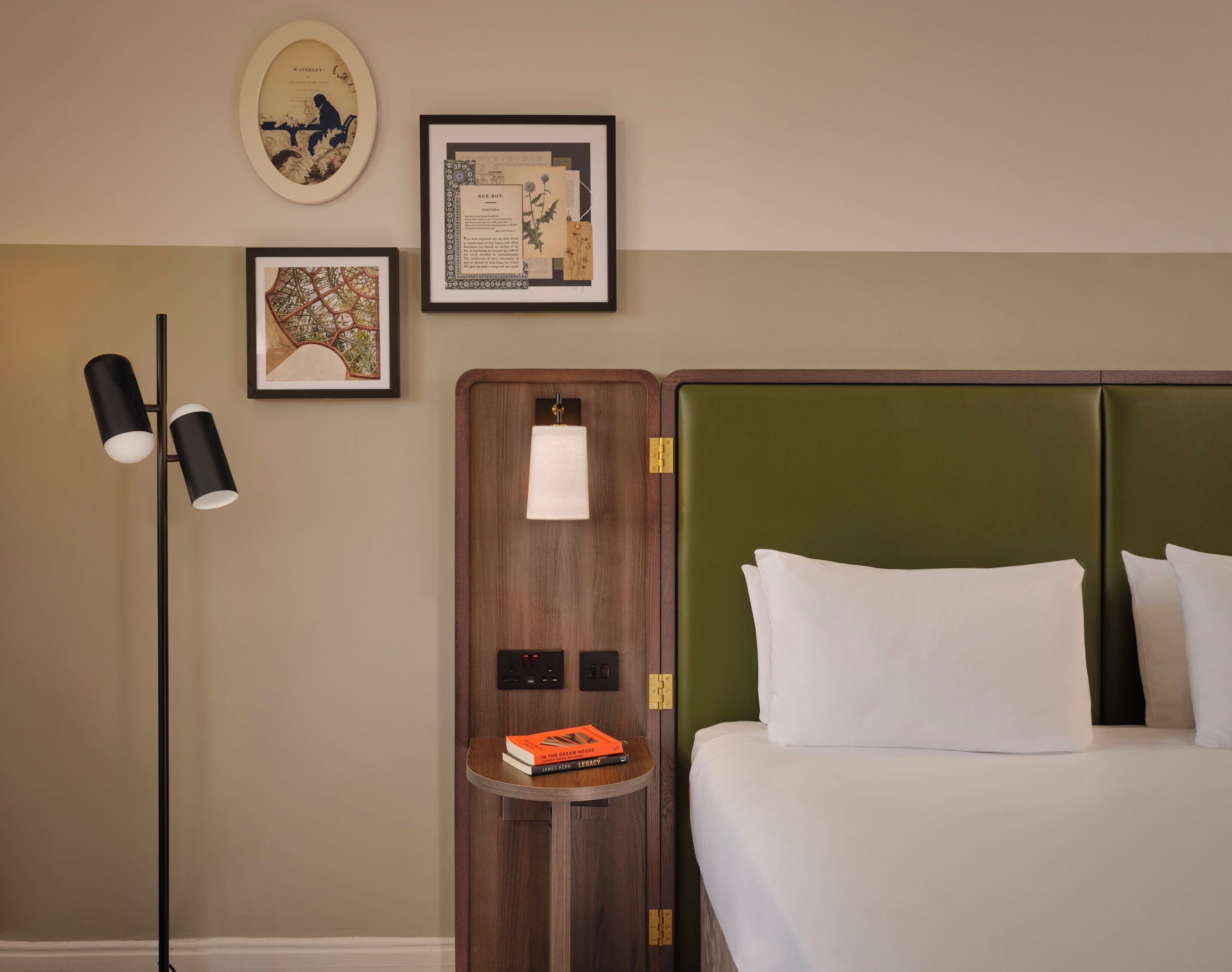 Een hotelkamer met een bed, een nachtkastje met een lamp en lijstjes aan de muur.