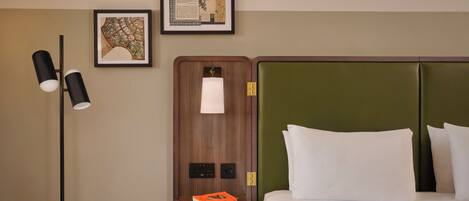 Een hotelkamer met een bed, een nachtkastje met een lamp en lijstjes aan de muur.