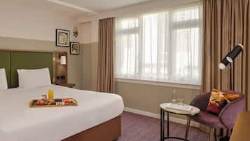 A hotel room with a large bed, a small table with a tray of breakfast, a chair with a cushion, and a window with curtains.