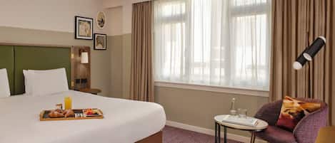 Una habitación de hotel con una cama grande, una mesita con una bandeja de desayuno, una silla con cojín y una ventana con cortinas.