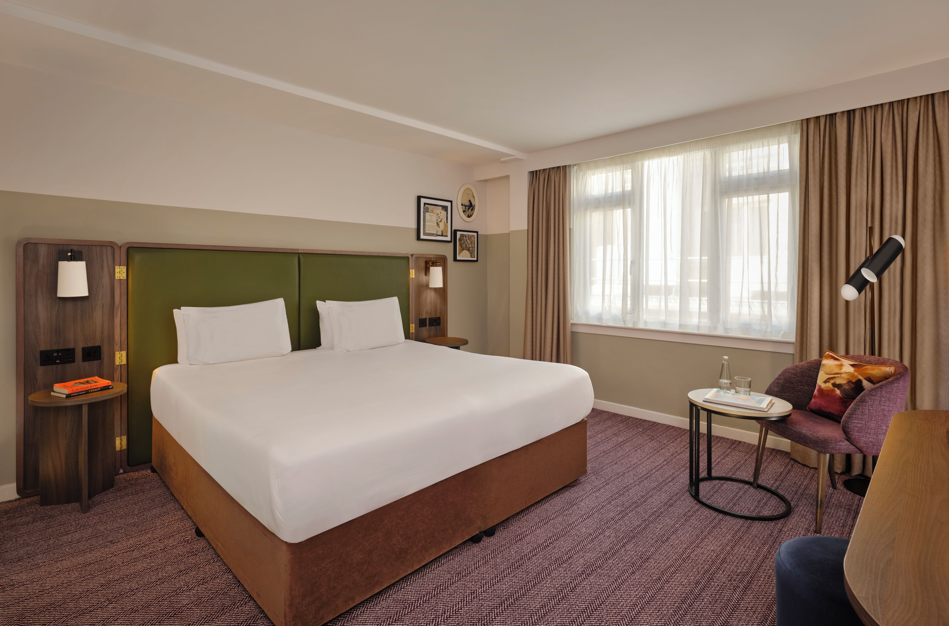 A hotel room with a large bed, a small table with a lamp, a chair, and a window with curtains.
