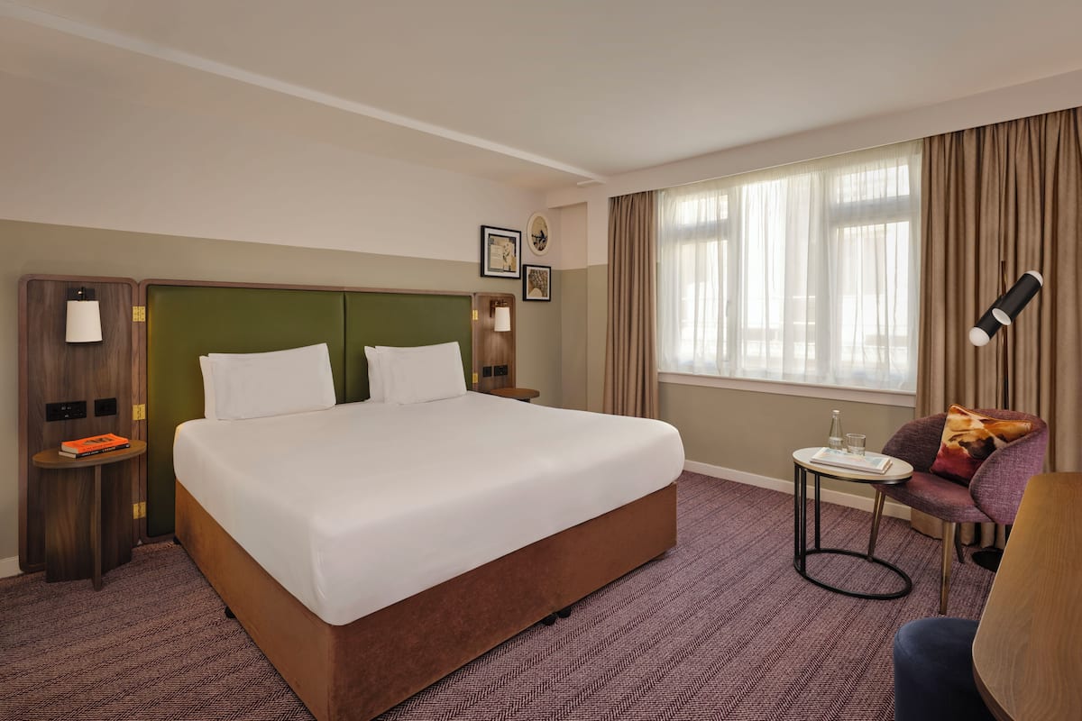 Habitación de hotel con una cama grande, una mesita de noche con lámpara, una silla y una ventana con cortinas.