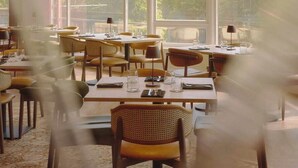 Un restaurante con amplios ventanales, mesas dispuestas con sillas y vistas a árboles y edificios.