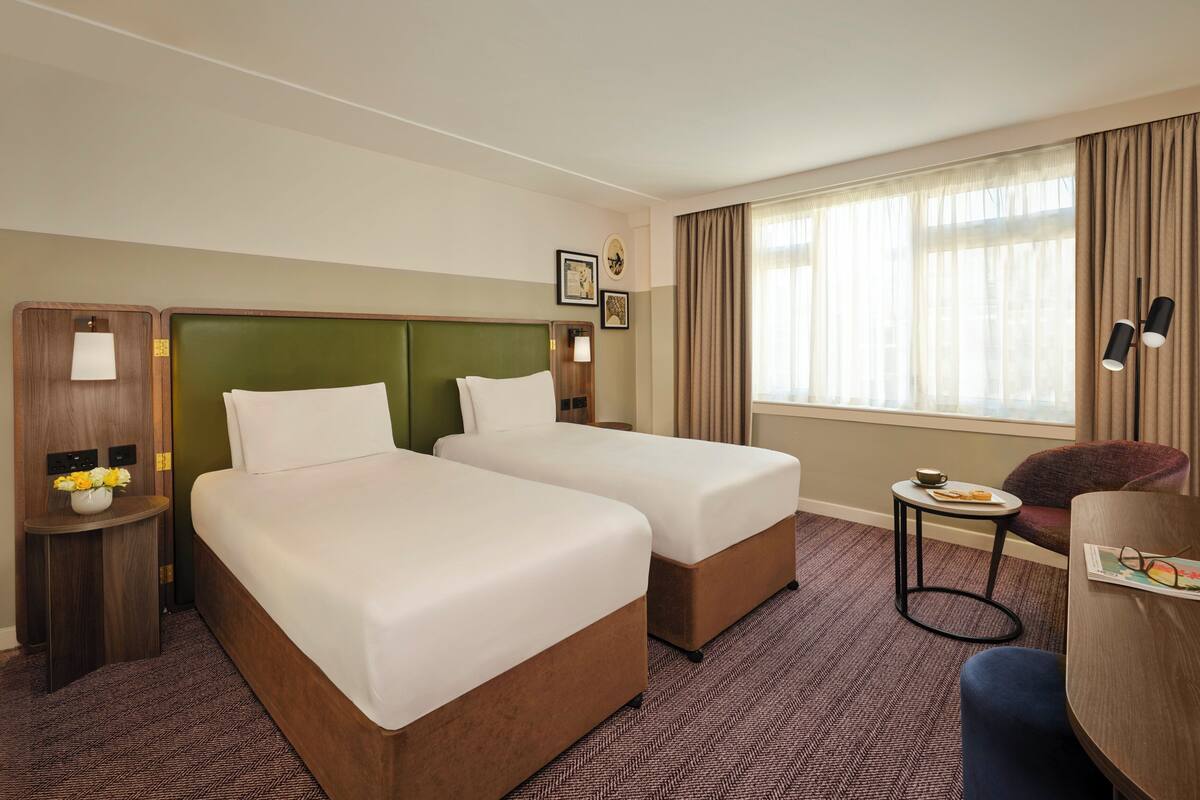 Habitación de hotel con dos camas, una mesita de noche con lámpara, una silla y una ventana con cortinas.