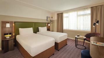 A hotel room with two beds, a small table with a lamp, a chair, and a window with curtains.
