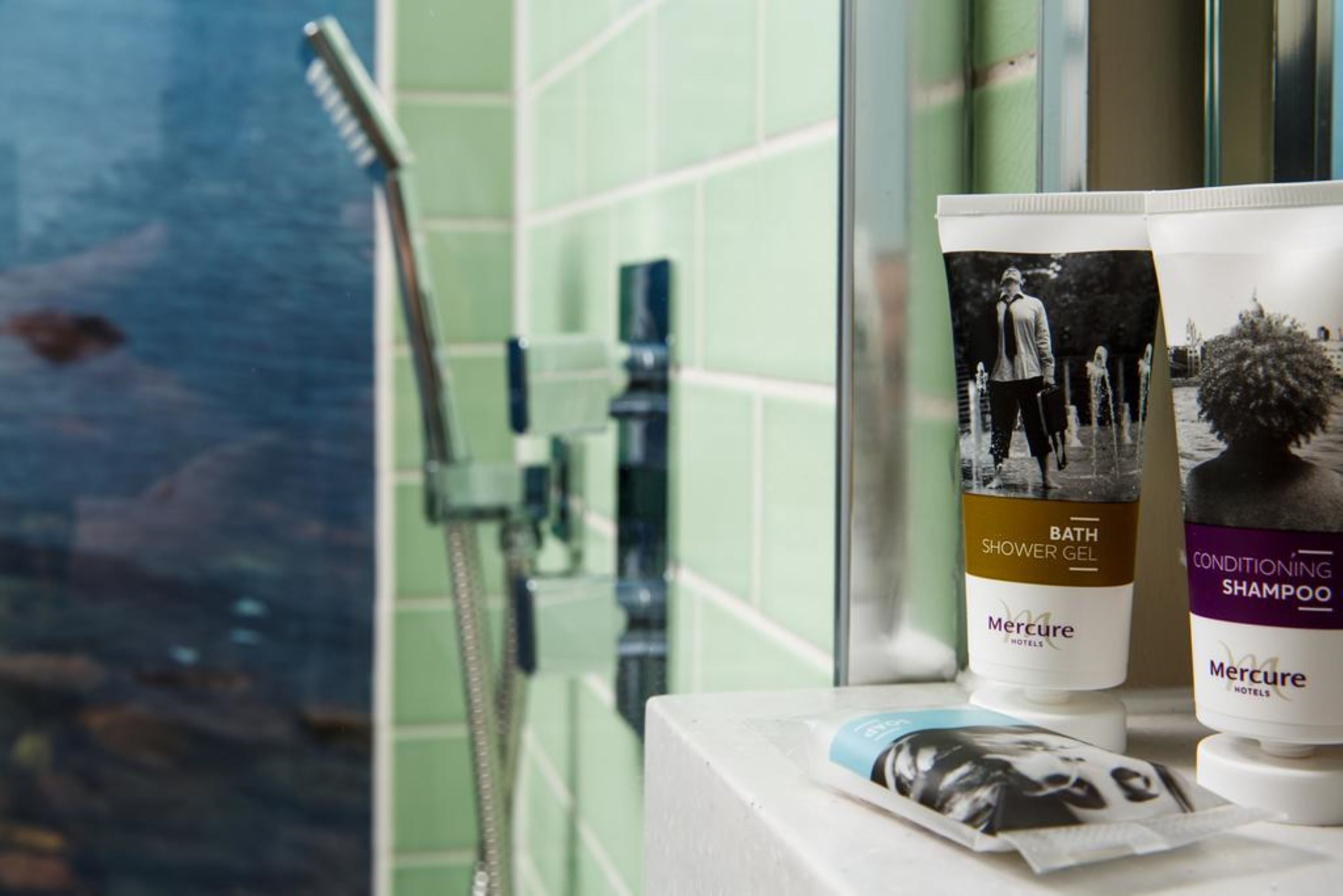 A bathroom with a shower, a bottle of Mercure hotel shower gel, and a bottle of Mercure hotel conditioning shampoo.
