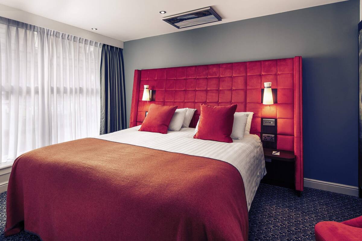 A hotel room with a large bed, red headboard, two bedside lamps, and a window with curtains.