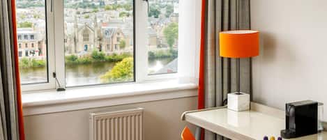 A hotel room with a view of a cityscape through large windows, a table with a lamp, a chair, and a radiator.