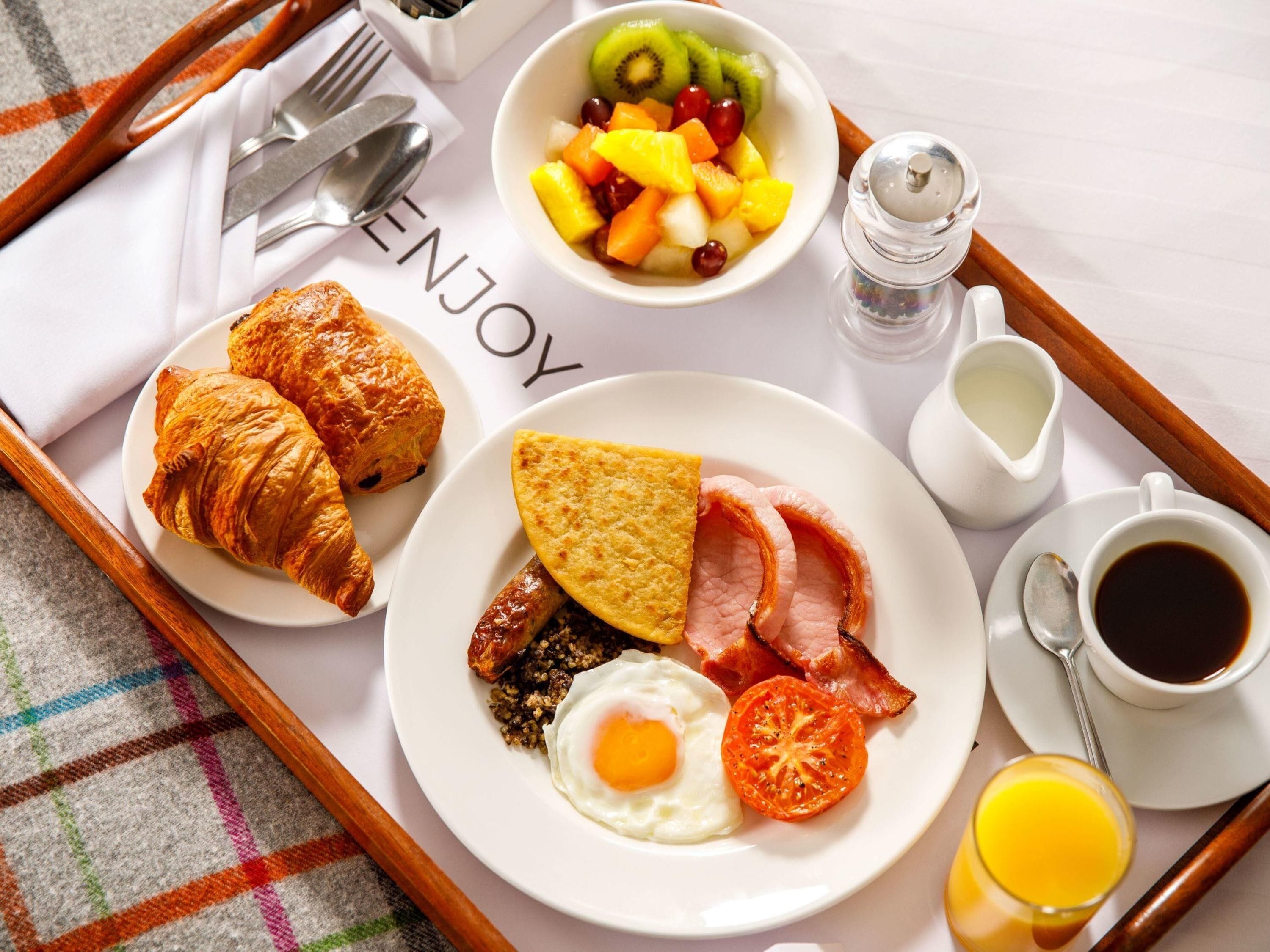 A breakfast tray with croissants, a fried egg, bacon, tomatoes, and a fruit bowl.