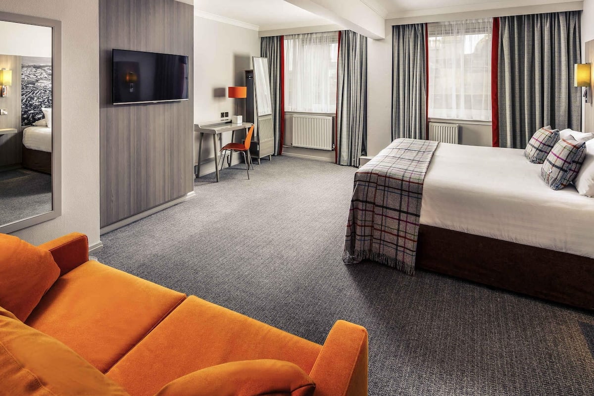 A hotel room with a bed, a desk, a chair, a TV, and a large window with curtains.