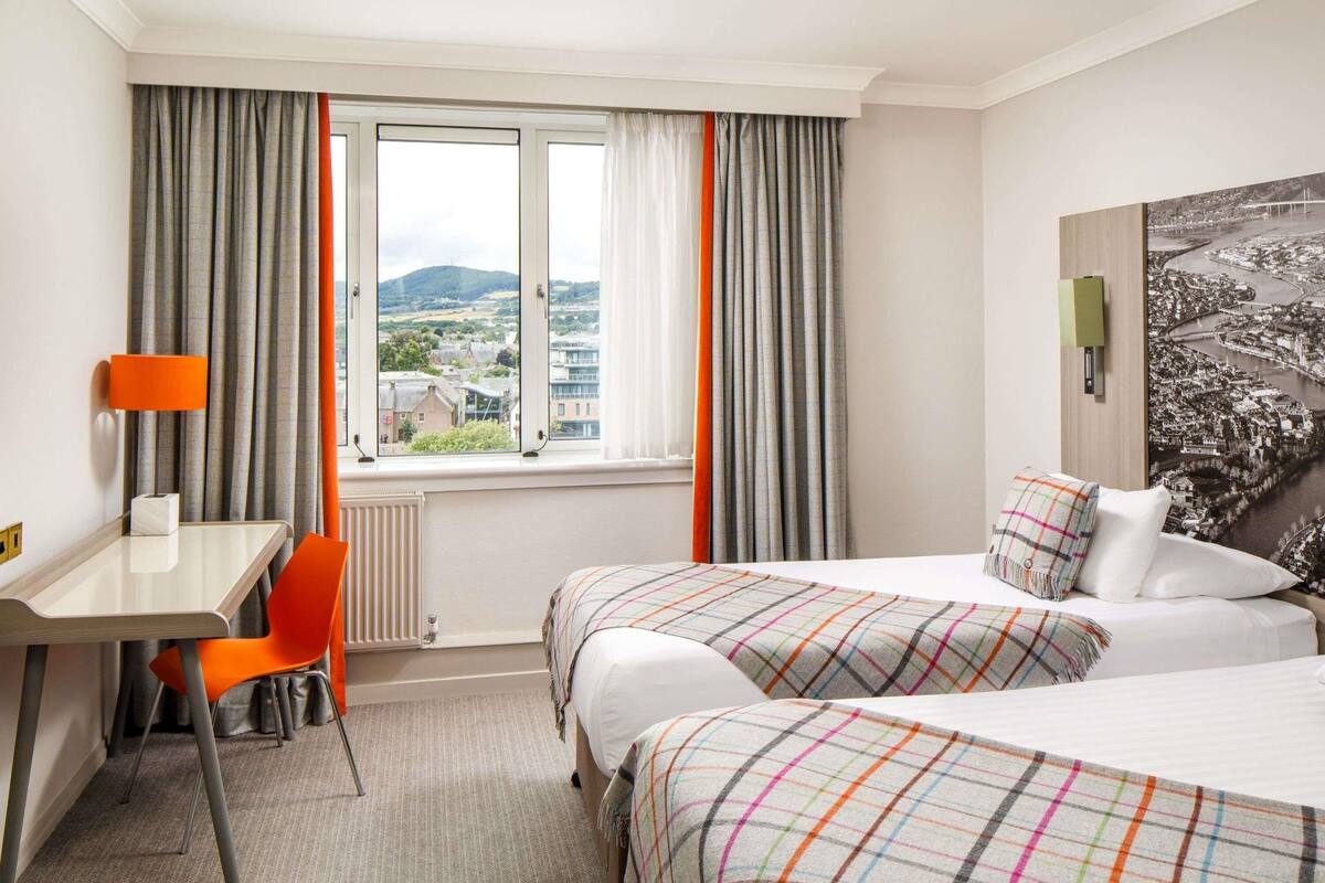 A hotel room with two beds, a desk, a chair, and a view of a cityscape.