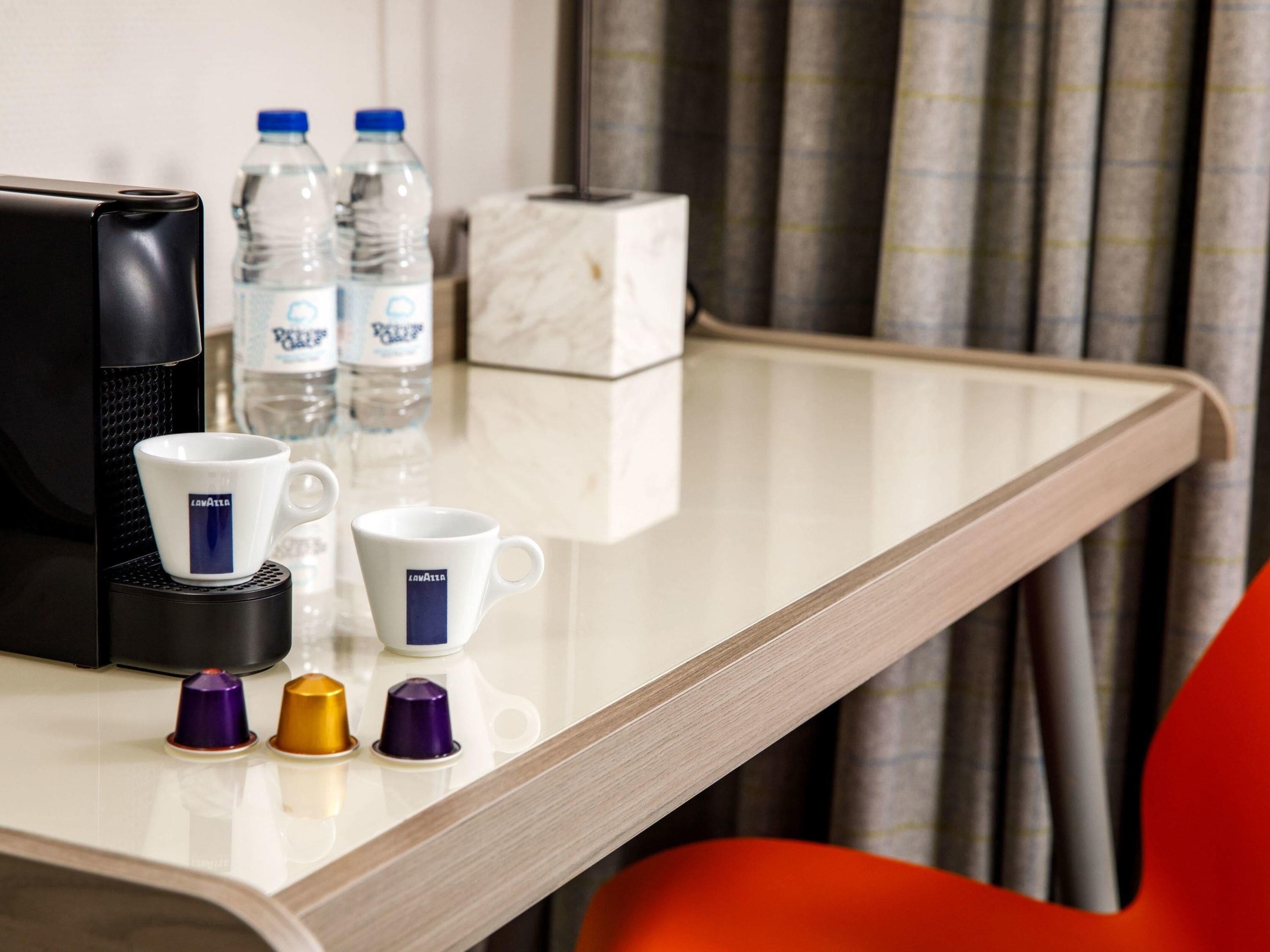 A hotel room with a coffee maker, two water bottles, a marble box, and a tray with coffee cups and capsules.