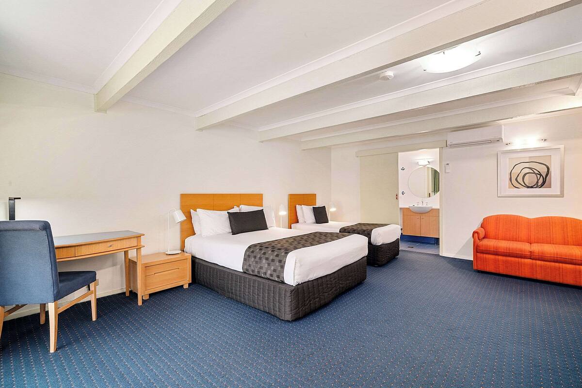 A hotel room with a large bed, a desk, a chair, a sofa, and a bathroom visible in the background.