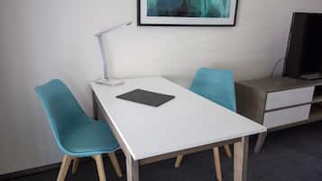 A modern office space with a white desk, two teal chairs, a desk lamp, a framed abstract painting, and a TV on a wooden stand.
