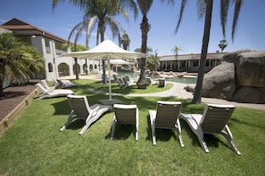 A poolside area with lounge chairs and umbrellas, surrounded by palm trees and a large rock formation. - Quality Resort Siesta (Lavington)