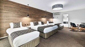 A modern hotel room with a wooden accent wall, multiple beds, a dining area, and a ceiling lamp.