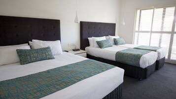 Habitación de hotel con dos camas, cada una con cabecera oscura y ropa de cama blanca. Se aprecia una ventana con persianas a la derecha.