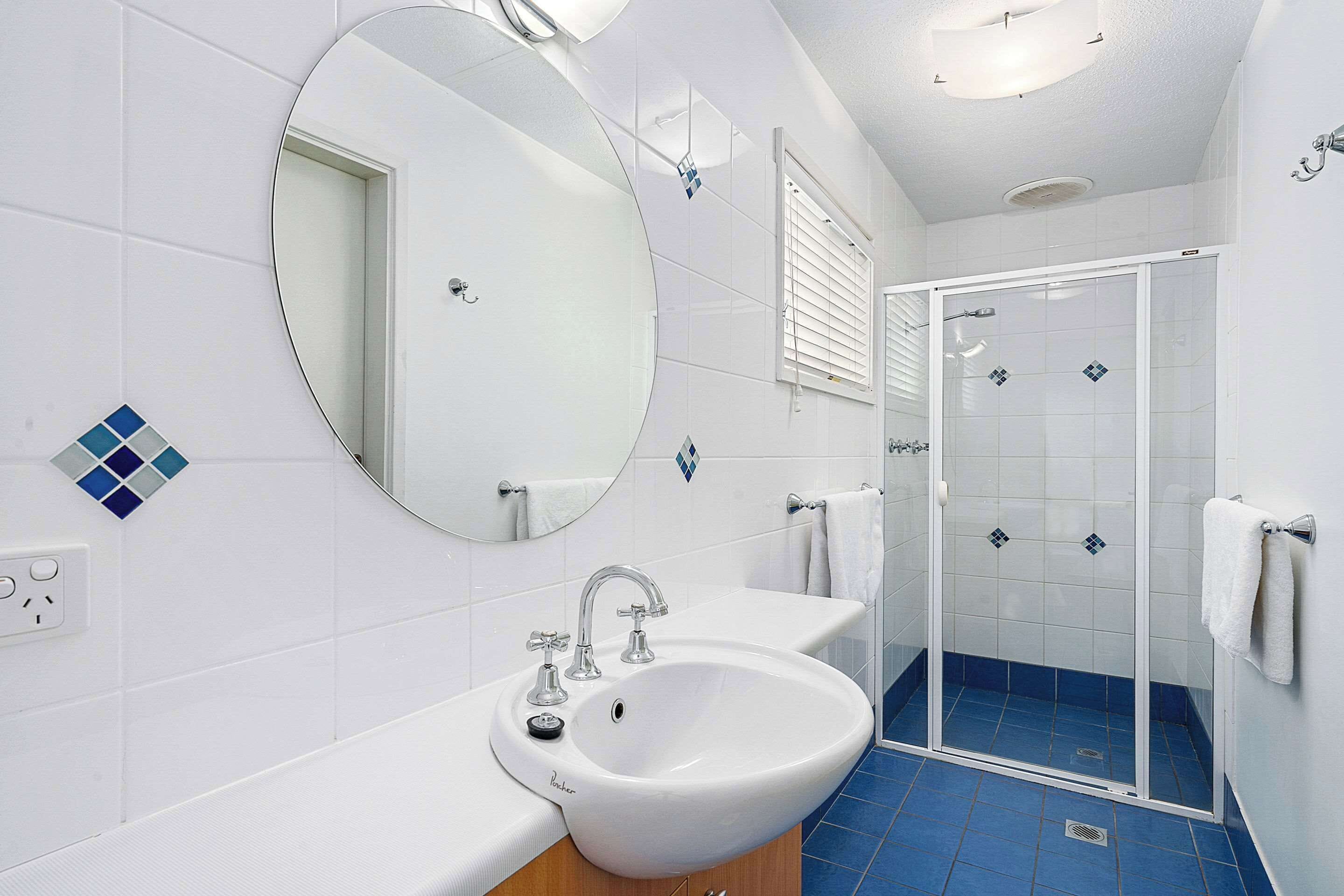 Un baño con un lavabo blanco, un espejo grande, ducha con puertas de cristal y suelo con azulejos azules.