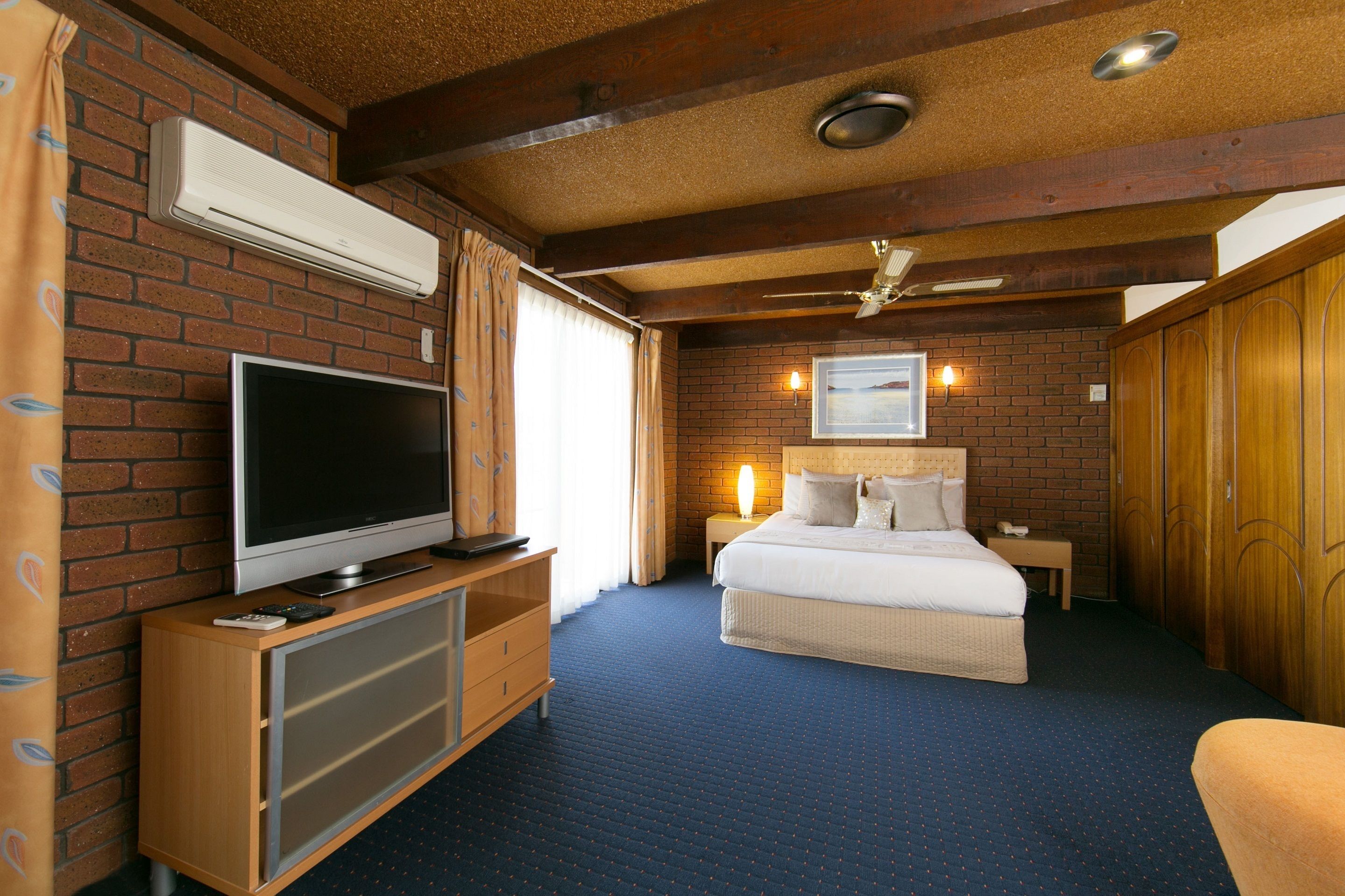 Habitación de hotel con pared de ladrillo, un televisor sobre un mueble de madera, una cama con ropa de cama blanca, una mesita de noche y un ventilador de techo.