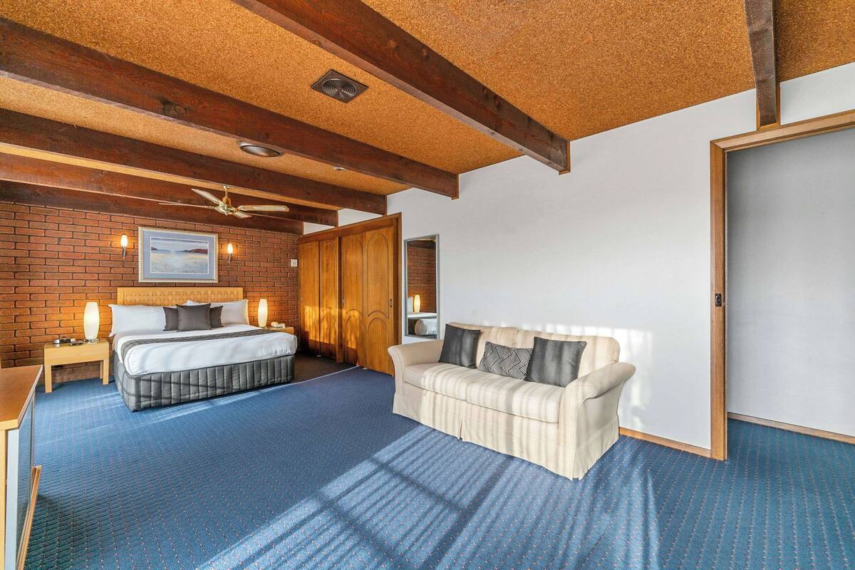 A bedroom with a bed, a sofa, a wooden ceiling, and a brick wall.