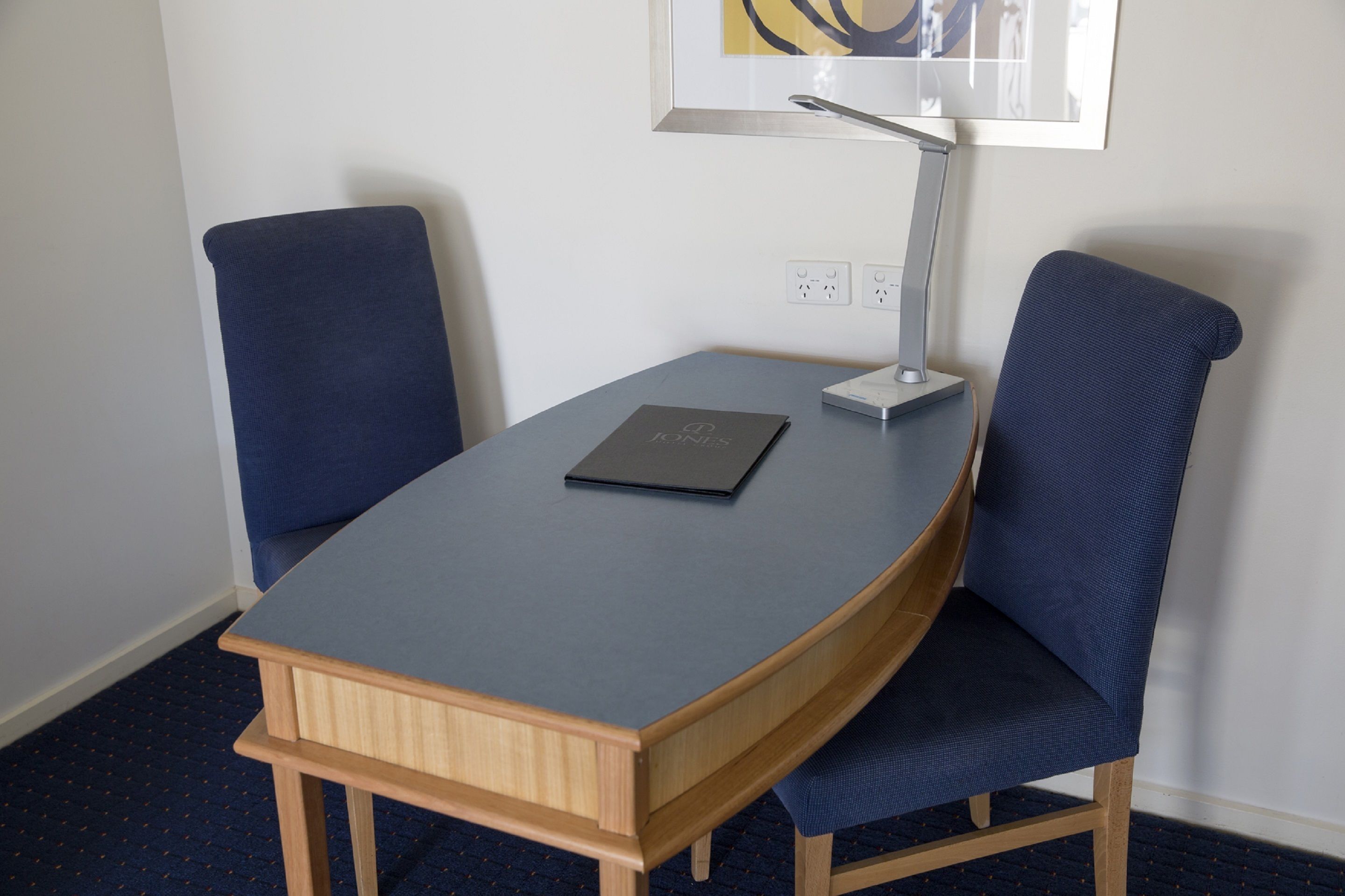 A small, modern room with a desk, two chairs, a lamp, and a framed picture on the wall.