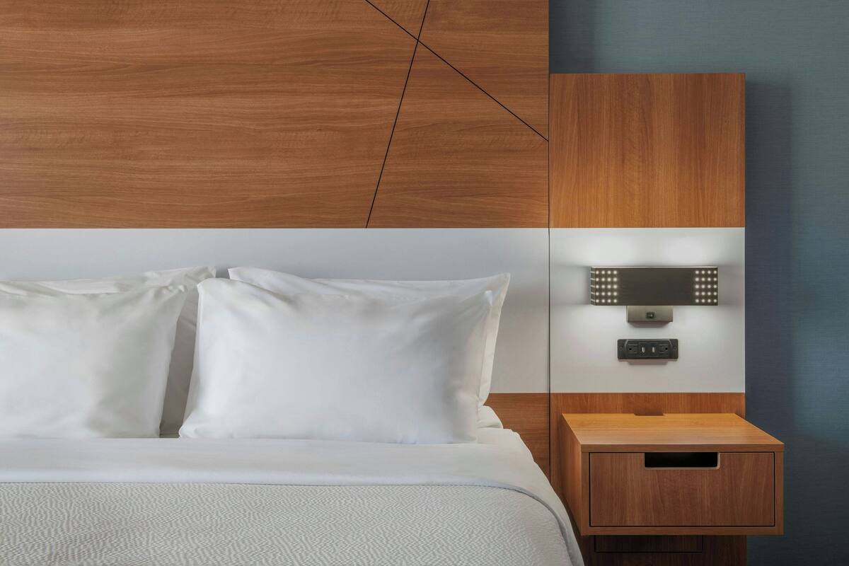 Ein modernes Hotelzimmer mit einem Holz-Kopfteil, einem Nachttisch mit integrierter Lampe und einem kleinen Holzbeistelltisch.