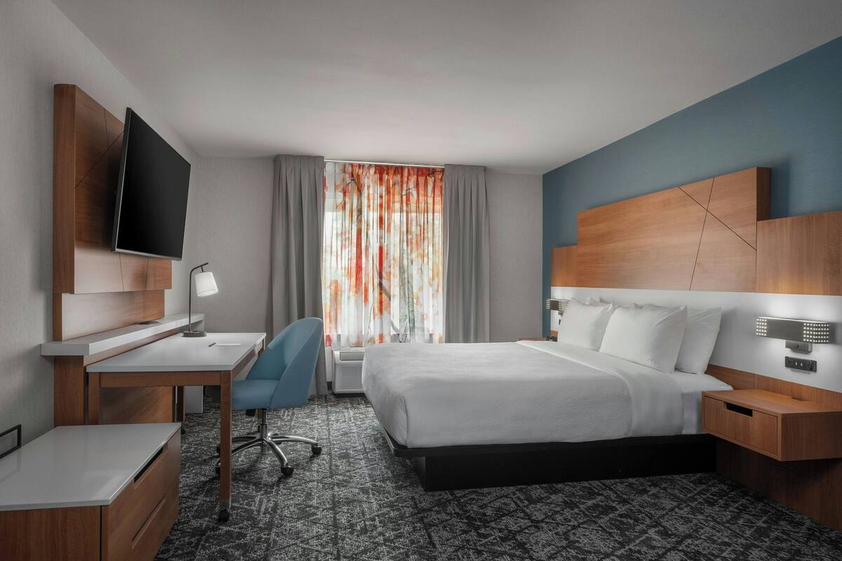 Ein Hotelzimmer mit Bett, Schreibtisch, Stuhl und einem an der Wand befestigten Fernseher.