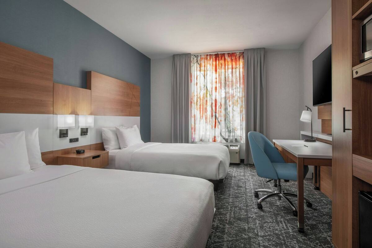 Ein Hotelzimmer mit zwei Betten, einem Schreibtisch, einem Stuhl, einem Fernseher und einem Fenster mit Vorhängen.