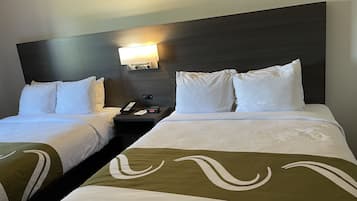 A hotel room with two beds, a nightstand with a phone and a digital clock, and a wall-mounted light fixture.